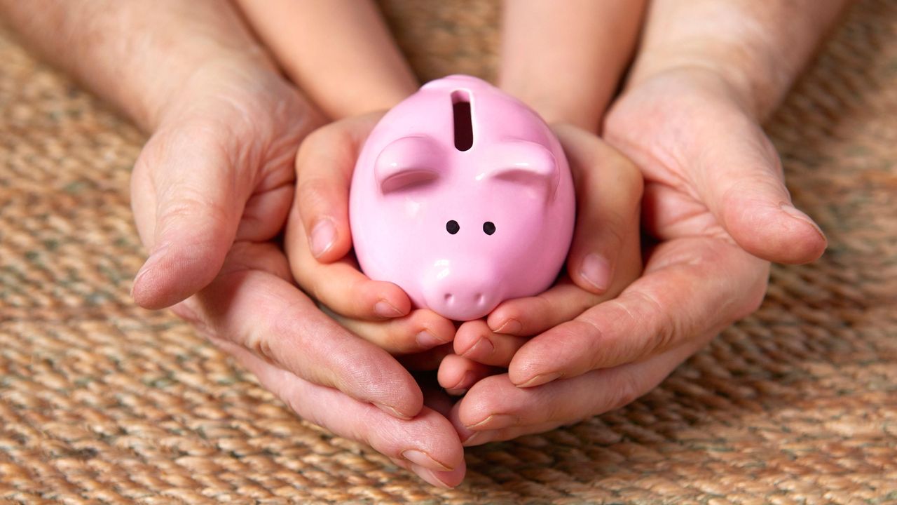 A parent&#039;s hands cradle a child&#039;s hands holding a piggy bank.