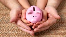 A parent's hands cradle a child's hands holding a piggy bank.