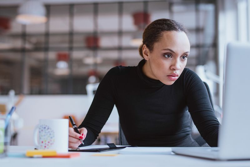 A young businesswoman looking at laptop while working at her desk. Female web designer taking notes from Internet.