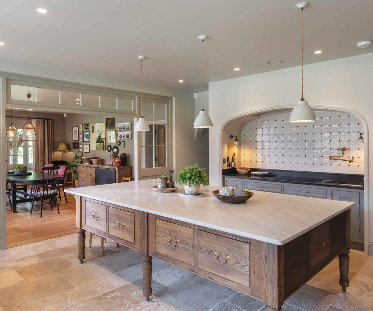 Open plan kitchen with wooden drawer island in the middle with hanging pendant lights above and a white marble work surface