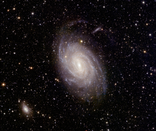 A gorgeous spiral galaxy is seen in the center of this image of space. In the background, there are lots of stars. Toward the bottom left of the scene, a way smaller hazy white blob.