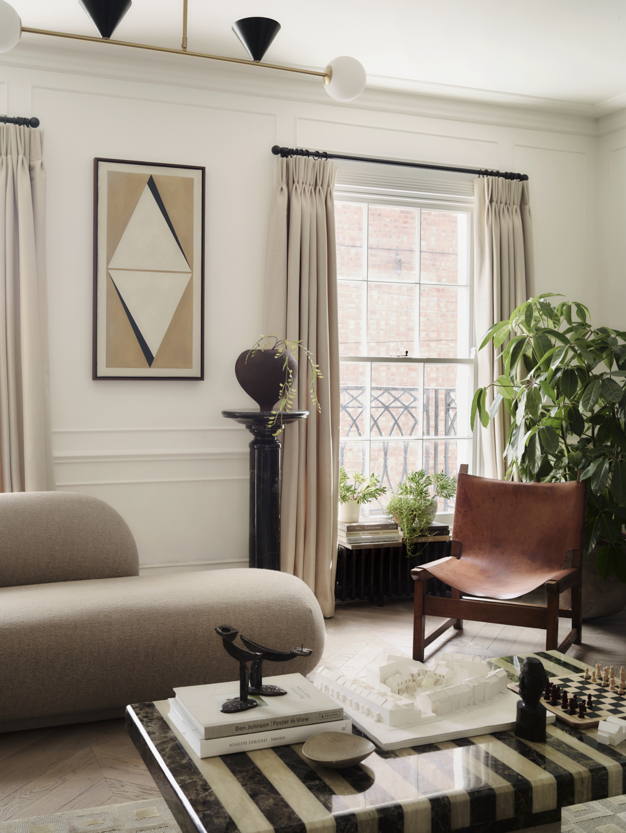 A formal, white living room with a window that has off-white curtains, a leather accent chair, a chaise lounge, a black pedestal with a vase, and a striped stone coffee table.