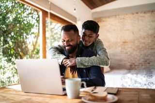 Stock photo of a young boy hugging his father as they look at a laptop screen and smile.