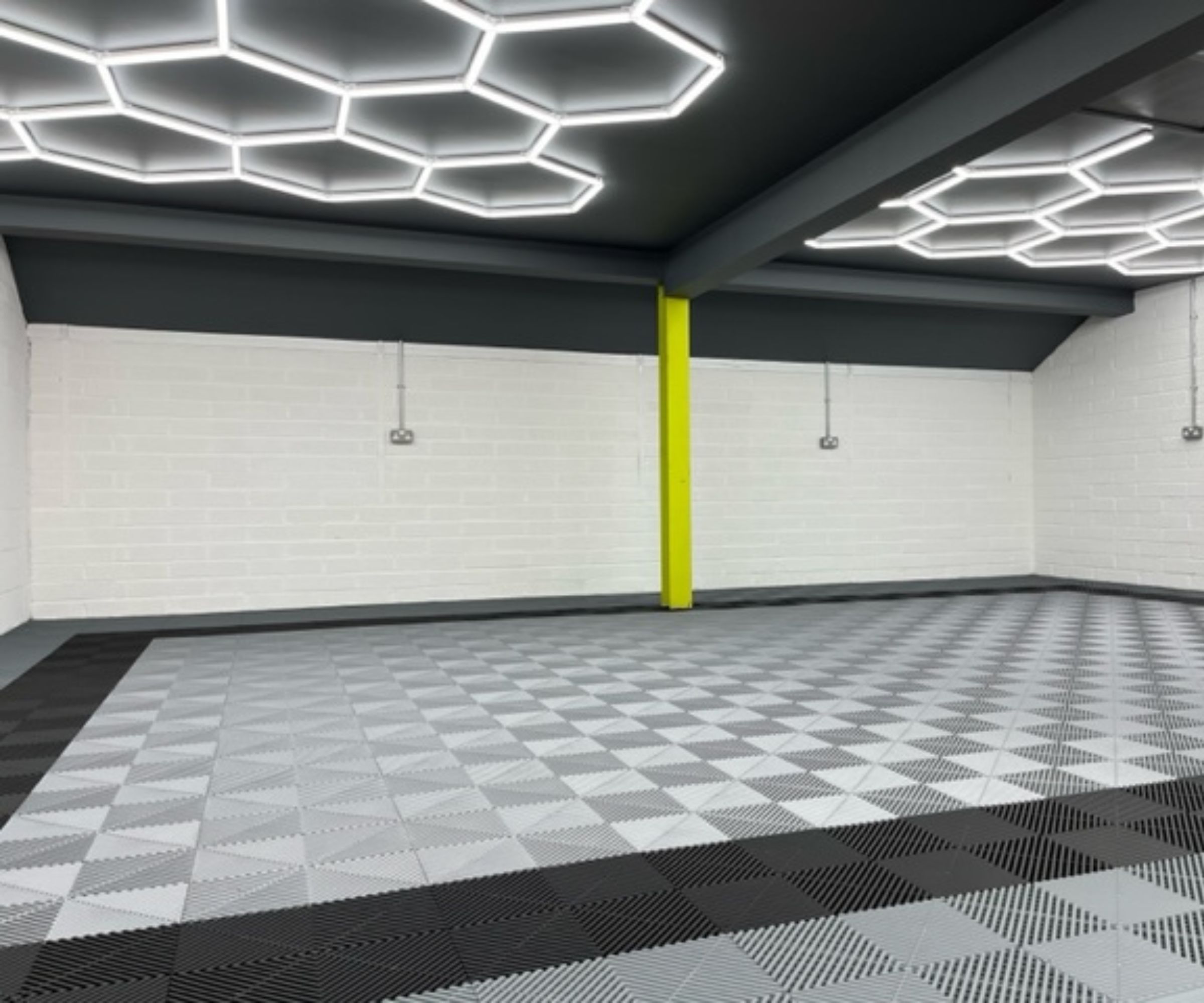 A large garage interior with bright hexagon-shaped LED ceiling lights, white brick walls, and a patterned grey and black tiled floor.