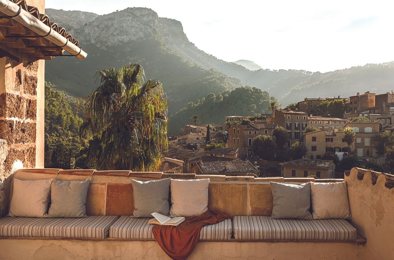 A rural retreat features colorful artworks and countryside views punctuated by sun-lit greenery and rocky buildings.