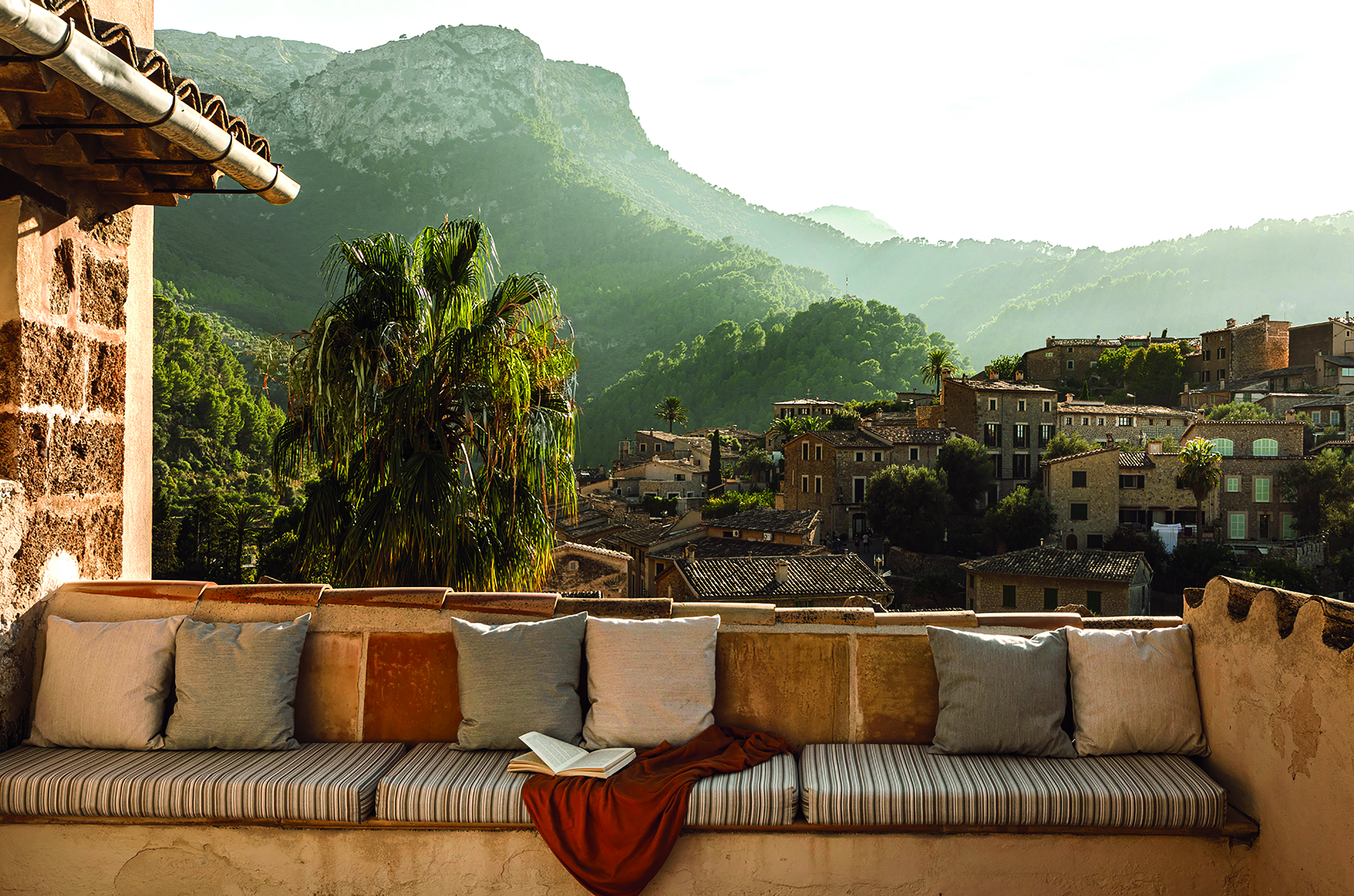 A rural retreat features colorful artworks and countryside views punctuated by sun-lit greenery and rocky buildings.