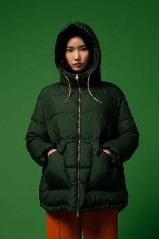 Female model wears Herno Globe A/W 2022 padded green coat, orange trousers against a green background