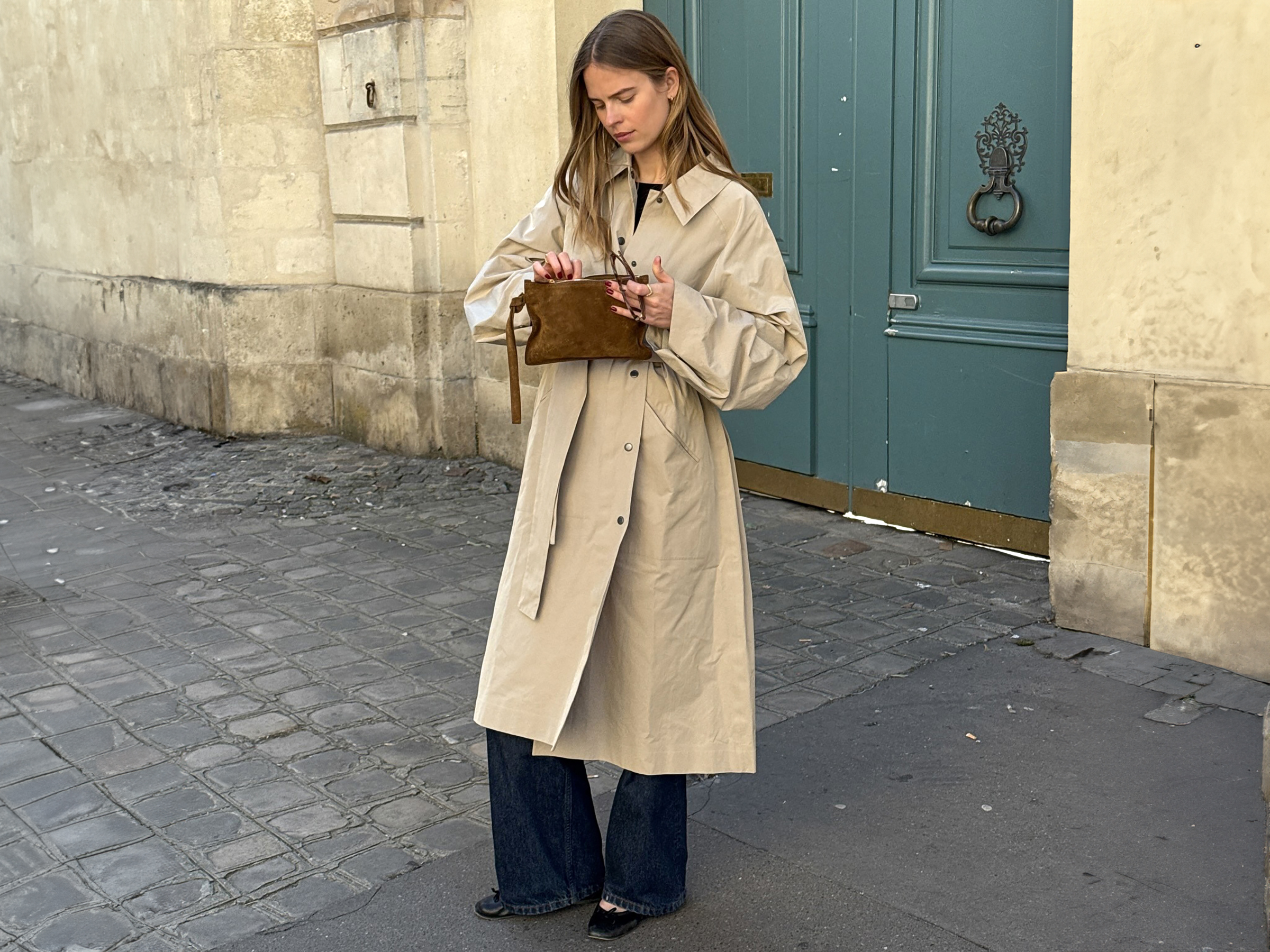 Scandi style influencer Clara Dyrhauge poses in Paris, France on a cobblestone street in front of an ornate teal green door wearing a calf-length trench coat, brown suede clutch bag, flared dark-wash jeans, and black ballet flats