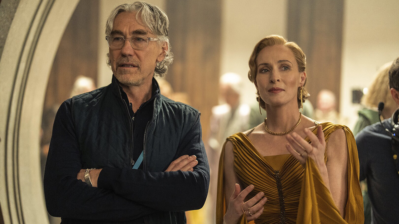 Still from the Star Wars TV show Andor. On the left is showrunner Tony Gilroy (older man with clear glasses, arms folded and wearing a blue jacket) on set. On the right next to him is Genevieve O’Reilly dressed in character wearing a beautiful gold dress and jewelry.