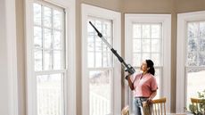 Person wearing pink shirt vacuuming window frame using the Shark Stratos Cordless, in dining area with three similar windows pictured to their left and right.