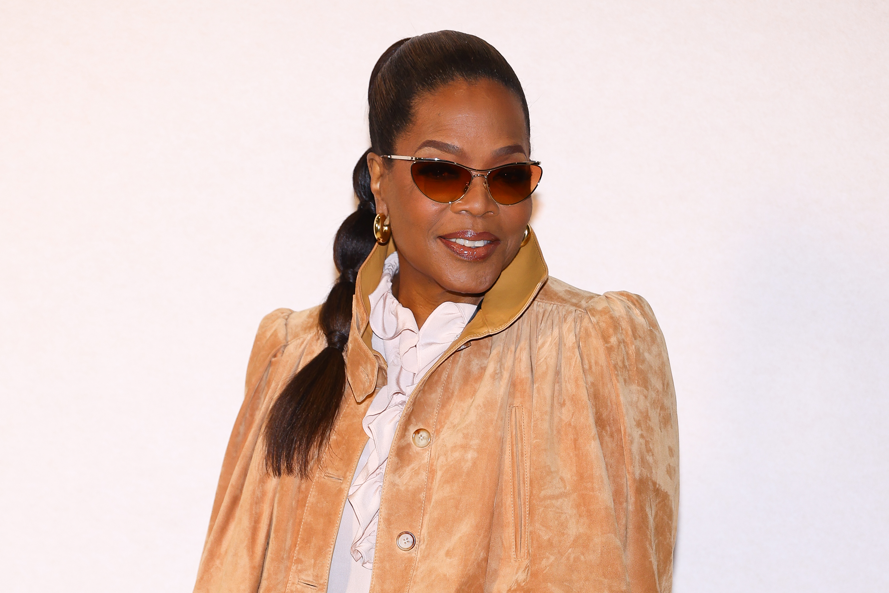 a black woman with a long ponytail wears a tan jacket and sunglasses while posing in front of a white background