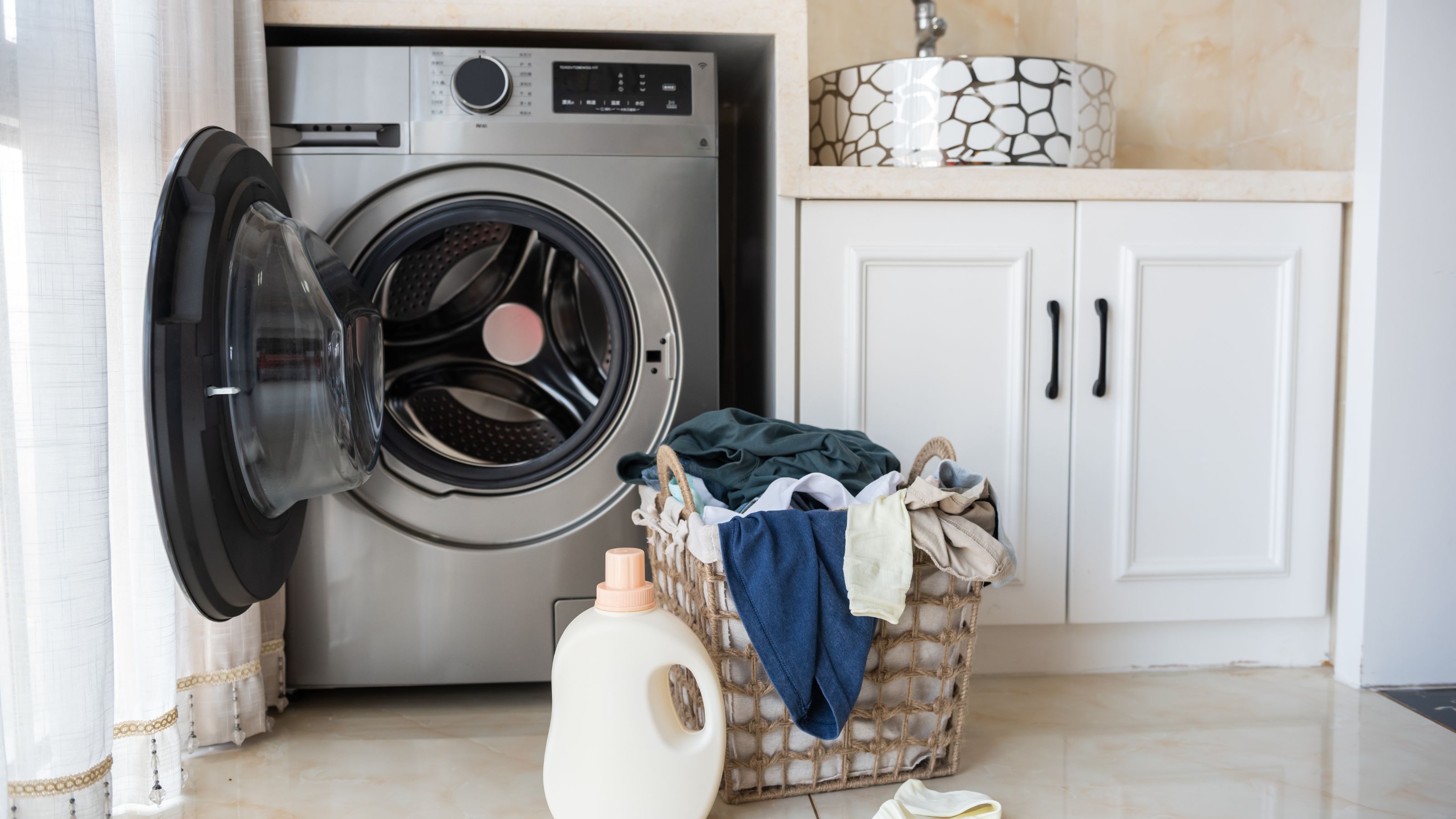 Open grey washing machine in utility room with basket of laundry and bottle of detergent on the floor in front 