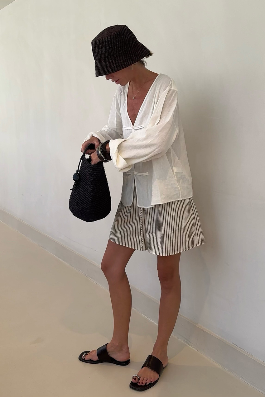 a woman wearing a black bucket hat, white longsleeve top, striped shorts, and black sandals