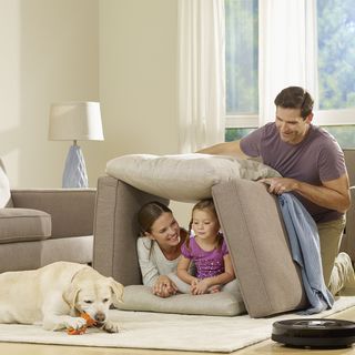 Family making pillow fort in a beige living room with a dog on the carpet and a robot vacuum cleaner close by