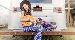 Bella Rayne sits on the back of a trailer with her Les Paul Goldtop, wearing tie-dyed patterned flares and cowboy boots.