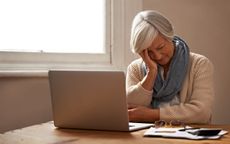 A woman with grey hair holds her head in her hand while sitting in front of a laptop.