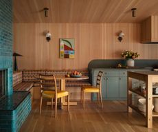 A wood-clad open-plan kitchen with wood floors, gray-green cabinets, banquette seating with a wood dining table and two mustard chairs. A teal brick fireplace. 