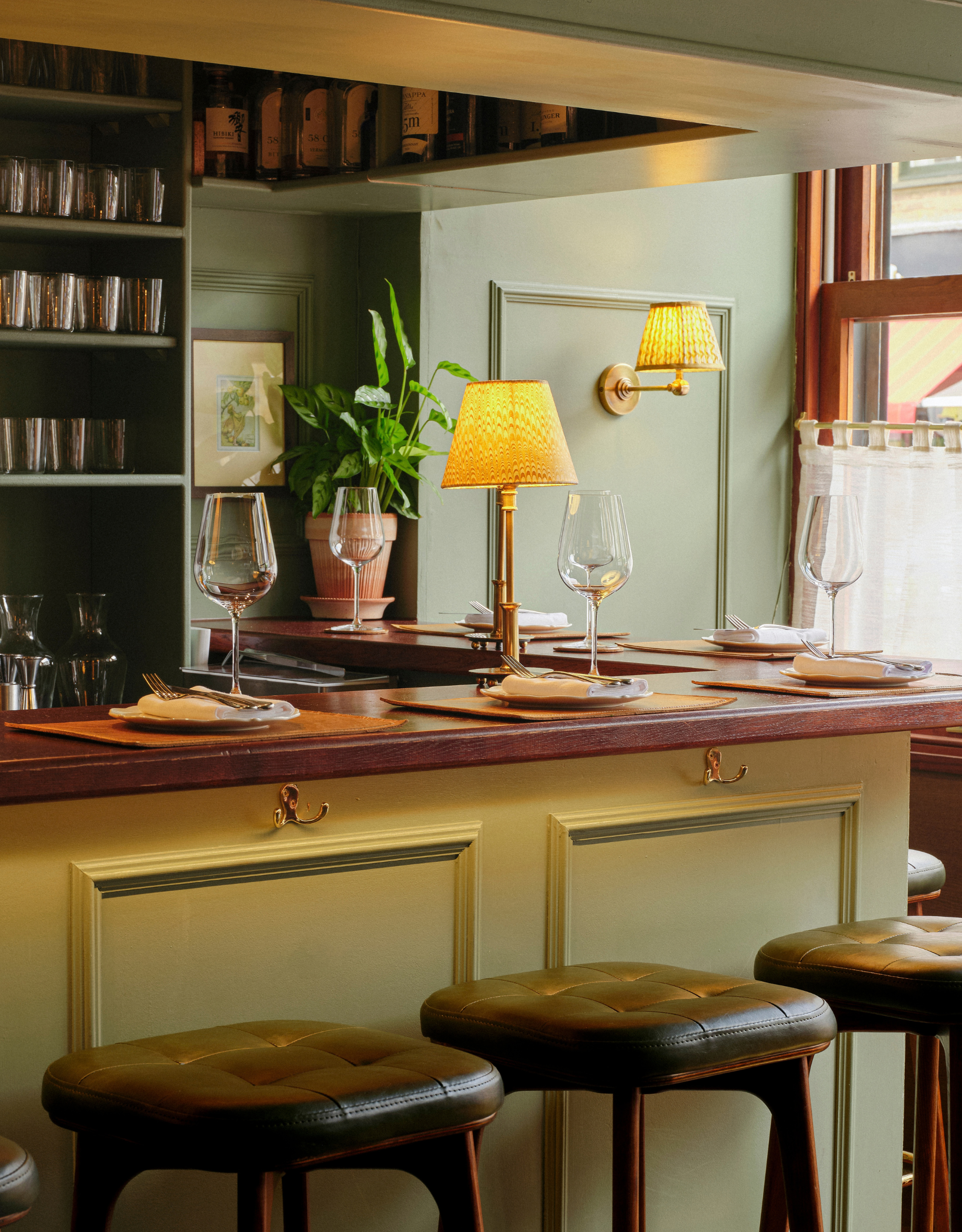 A sage green restaurant with a wood-top bar set with placemats, plates, cutlery, and wine glasses