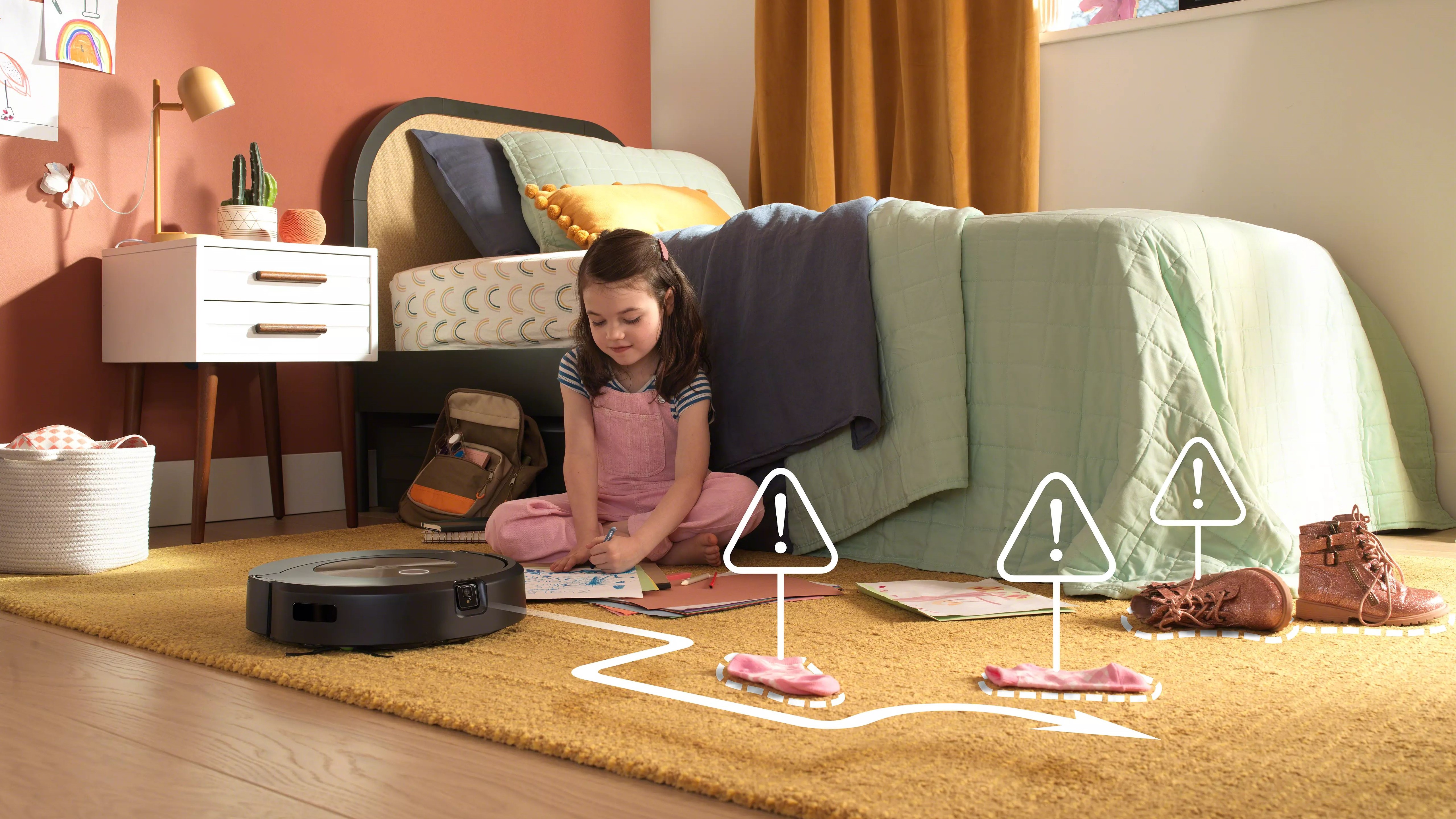 A Roomba j9+ navigating a child's room with socks and boots strewn across it - and a little girl drawing on the floor.