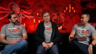 Three Diablo 4 developers sitting on a couch with shirts that say DAMAGE ON TUESDAYS in front of a red-lit background