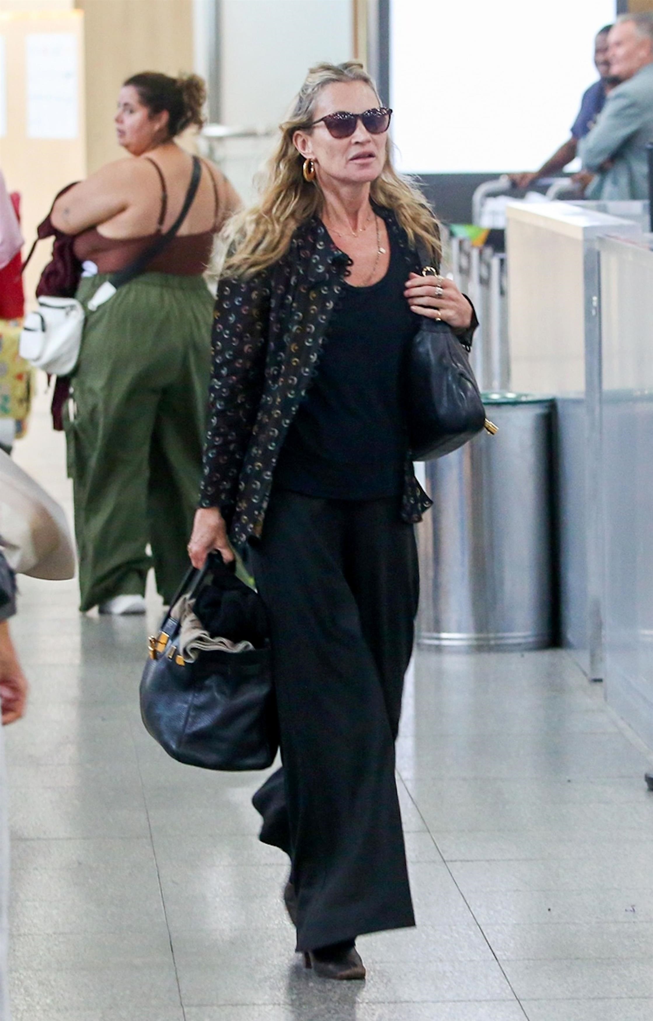 Kate Moss walks through the airport wearing black wide-leg jeans with a black vest top, a printed shirt and heeled boots.