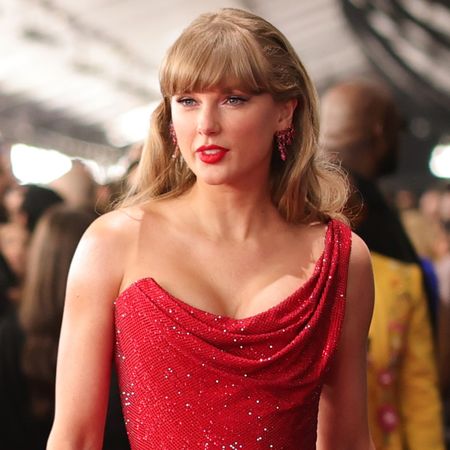 Taylor Swift wearing a red sequined one shoulder minidress walking the Grammys 2025 red carpet with a crowd of photographers and people blurred behind her