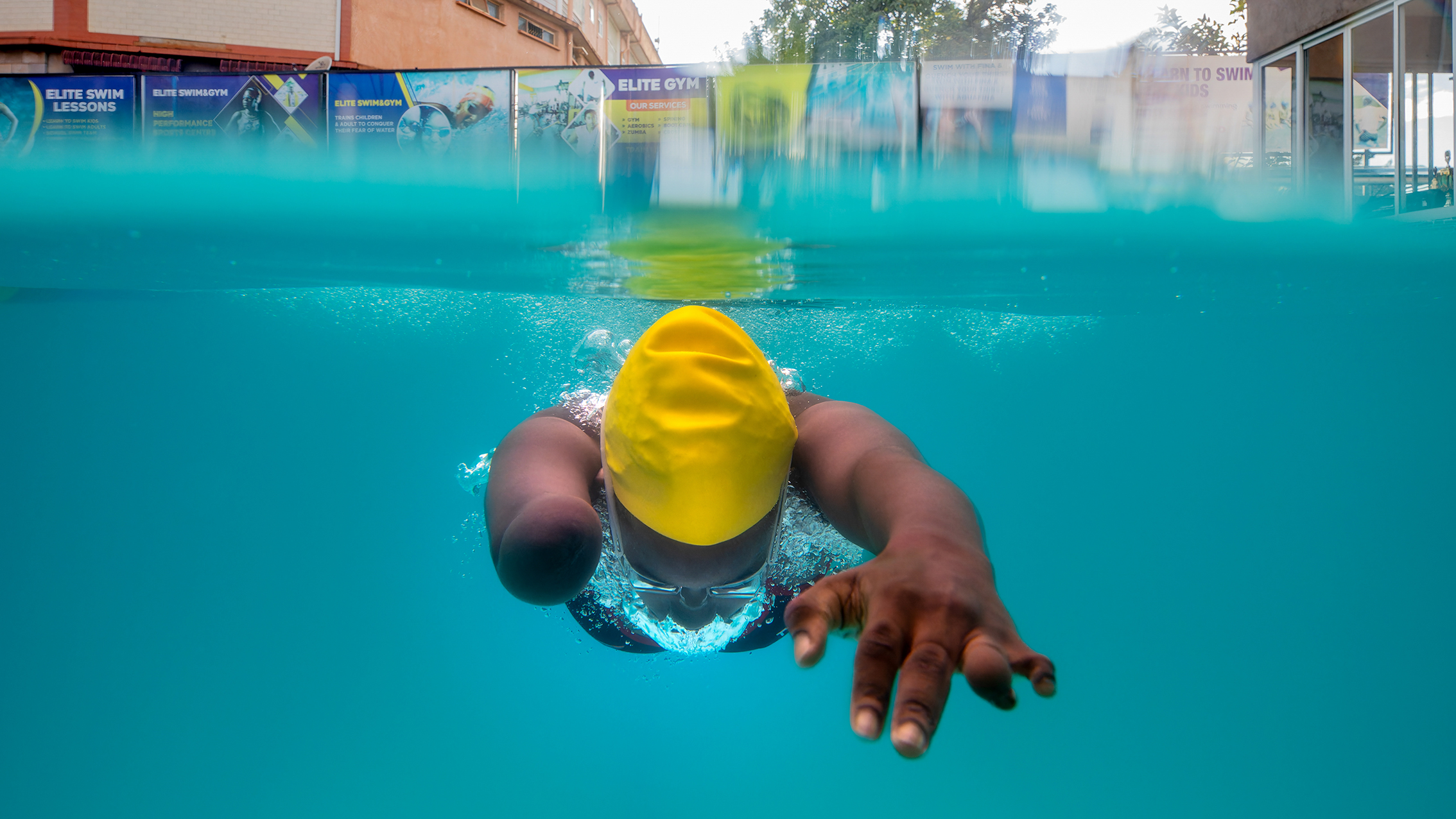 A swimmer in a yellow cap glides through clear blue water, one arm extended forward and bubbles trailing behind