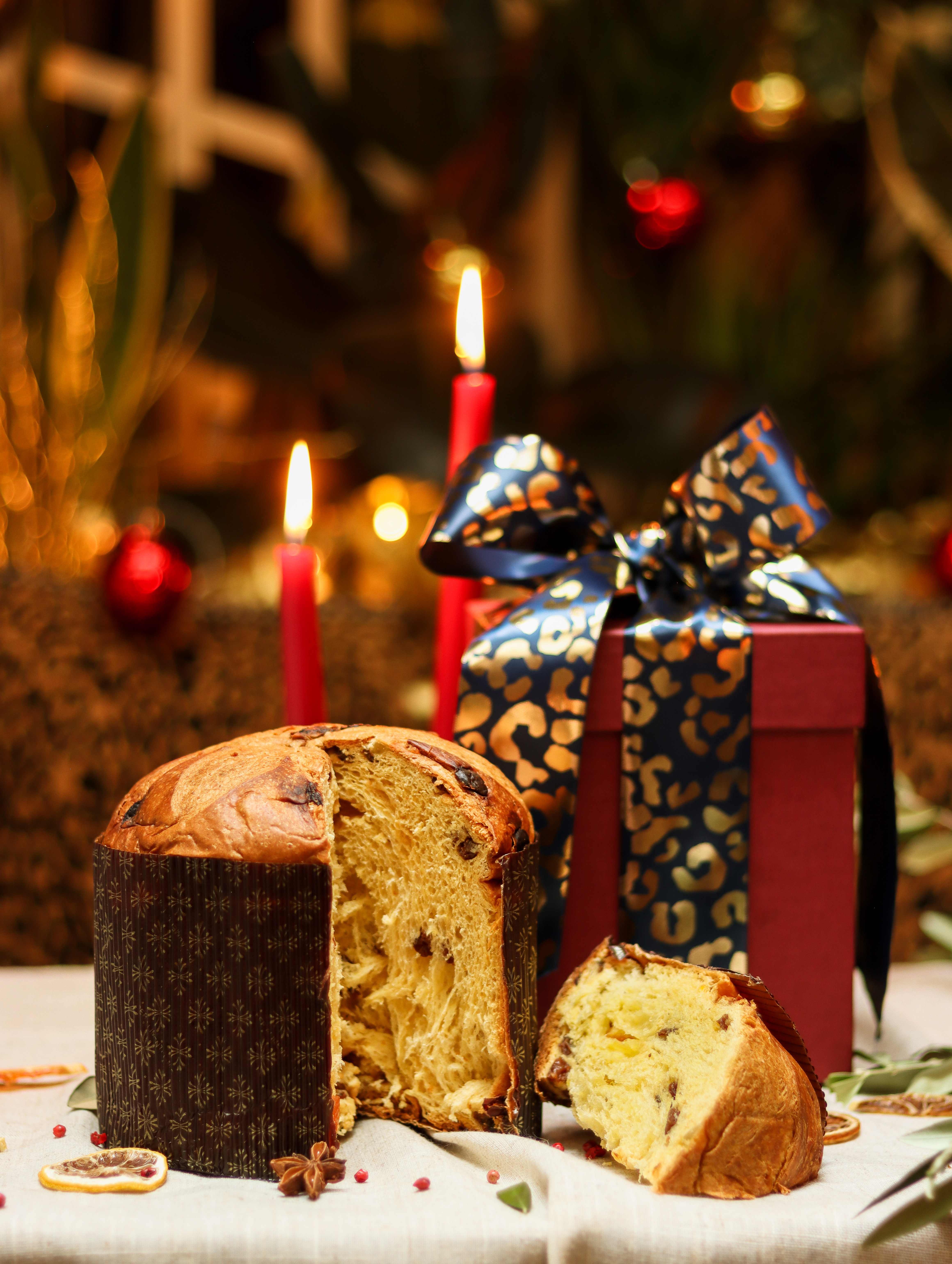 A candy peel and raisins-filled cake wrapped in a brown foil sits next to some wrapped presents and before lit red candles in a Christmassy setting.