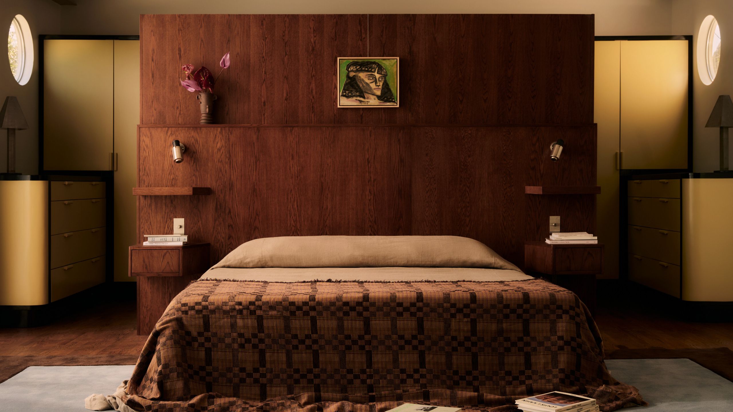 A modern bedroom with a large wooden headboard with built-in nightstands and shelving, with portholes on either side of the bed which is draped with a patterned bedspread that puddles on the floor