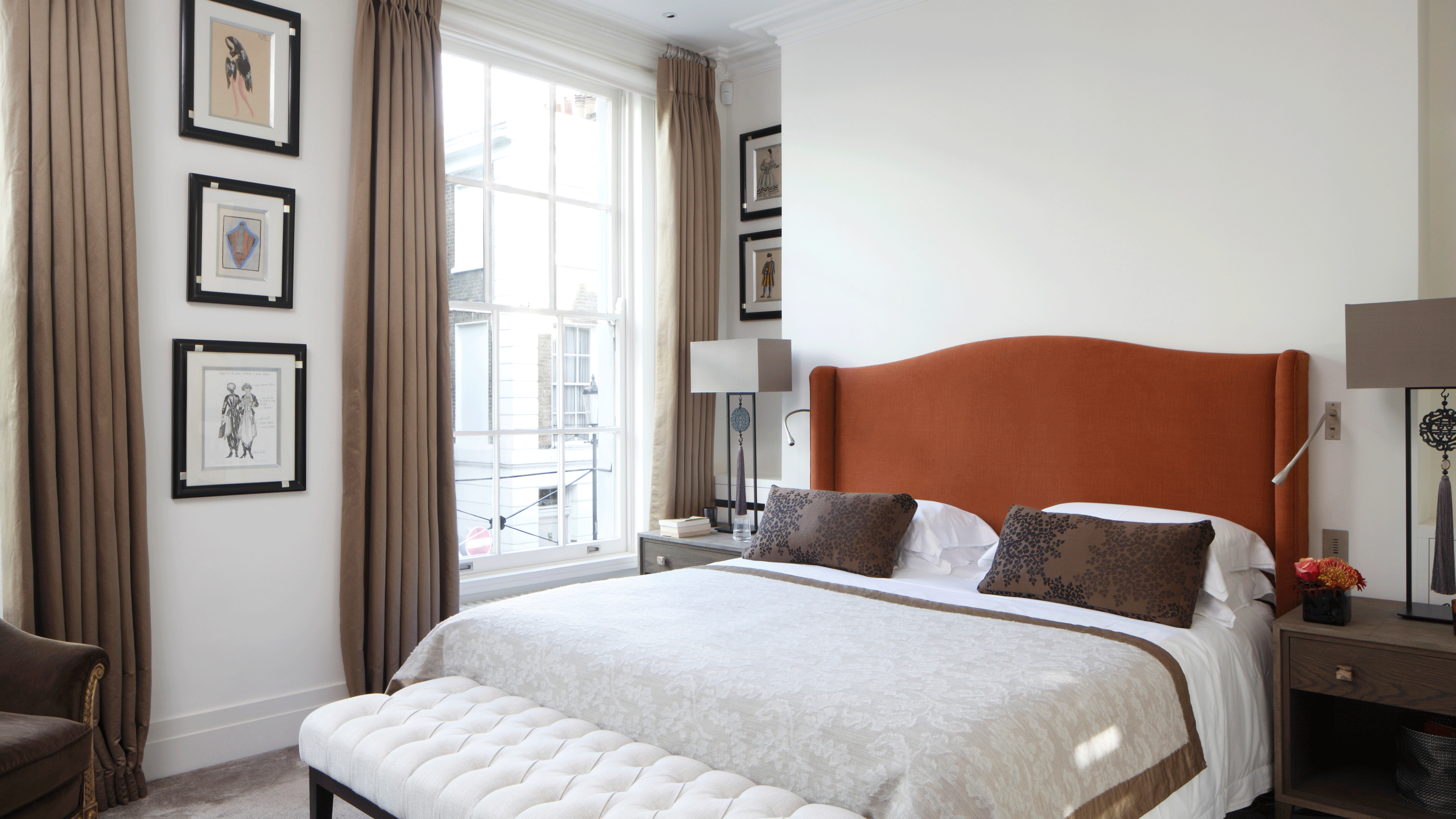 A white bedroom with large sash windows surrounded by brown floor length curtains. A bed with orange headboard and white bedding sit in front.