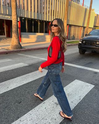 Woman on street wears red jumper, blue jeans and black sandals