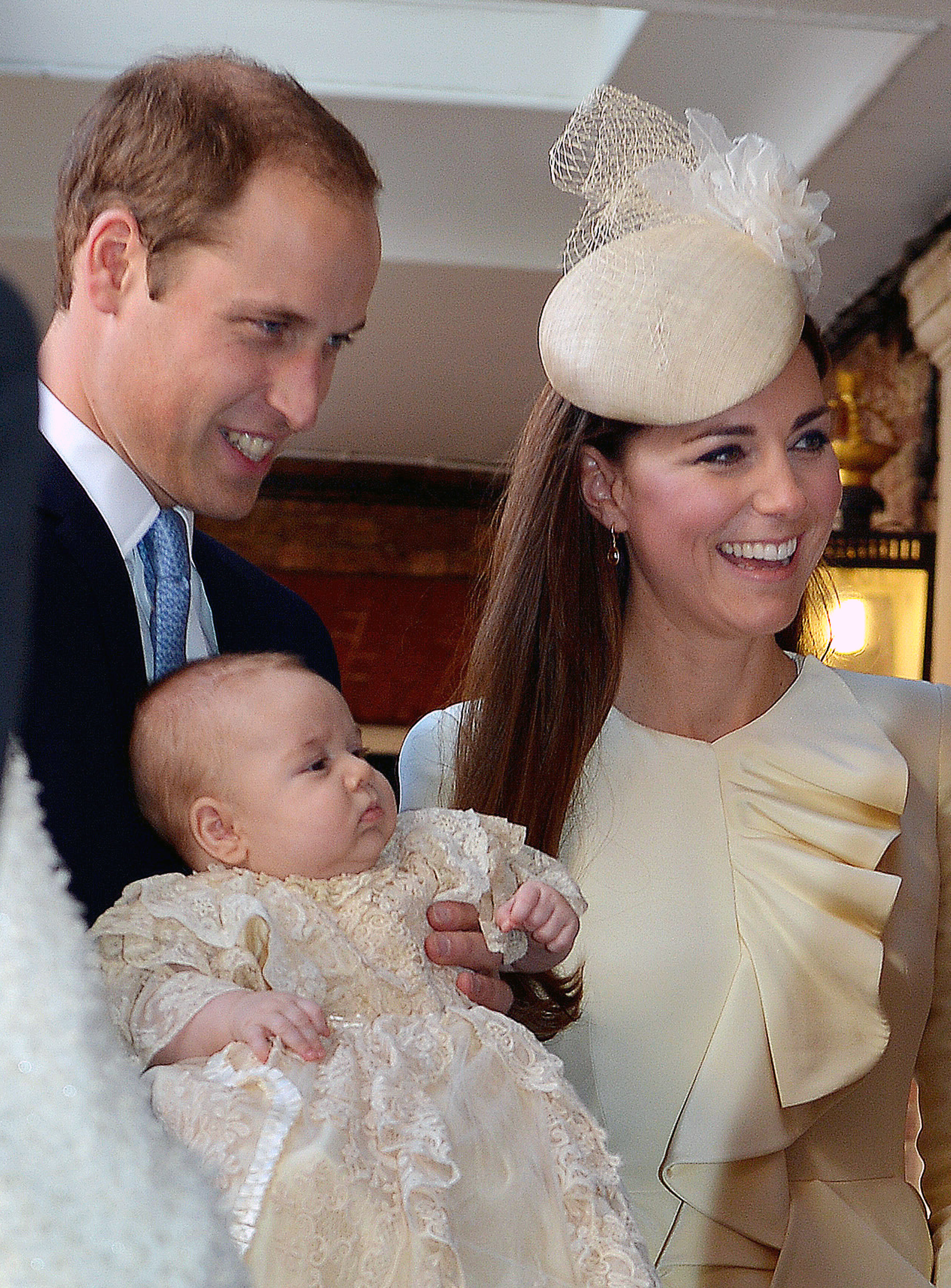 Kate Middleton wearing a cream hat and ruffled coat standing next to Prince William, who is holding Prince George, in a christening gown