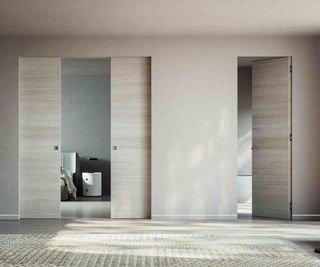 A light-filled landing space painted in a warm neutral colour, with wood flooring and a large woven rug. There are two doors — one a standard opening doors; the other, a set of pocket sliding doors by ECLISSE, opening to a bedroom.