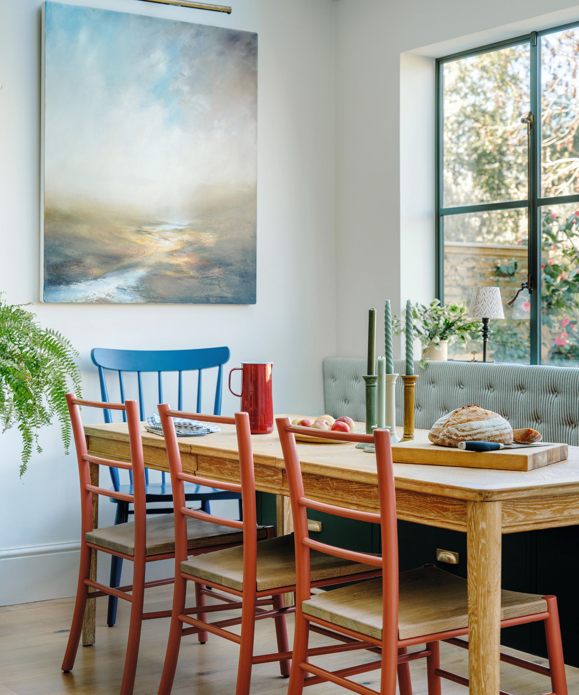 a dining nook with white walls and a large window out to the garden with a green striped banquette built beneath. A rustic wooden table is paired with red chairs