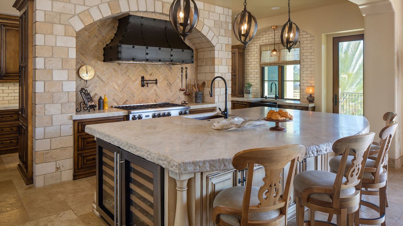 We love how gorgeous French kitchen ideas are. Here one with three iron pendant lights, a stone countertop with a sink integrated in it and a wine fridge, with an oven with a black studded hood to the back of it