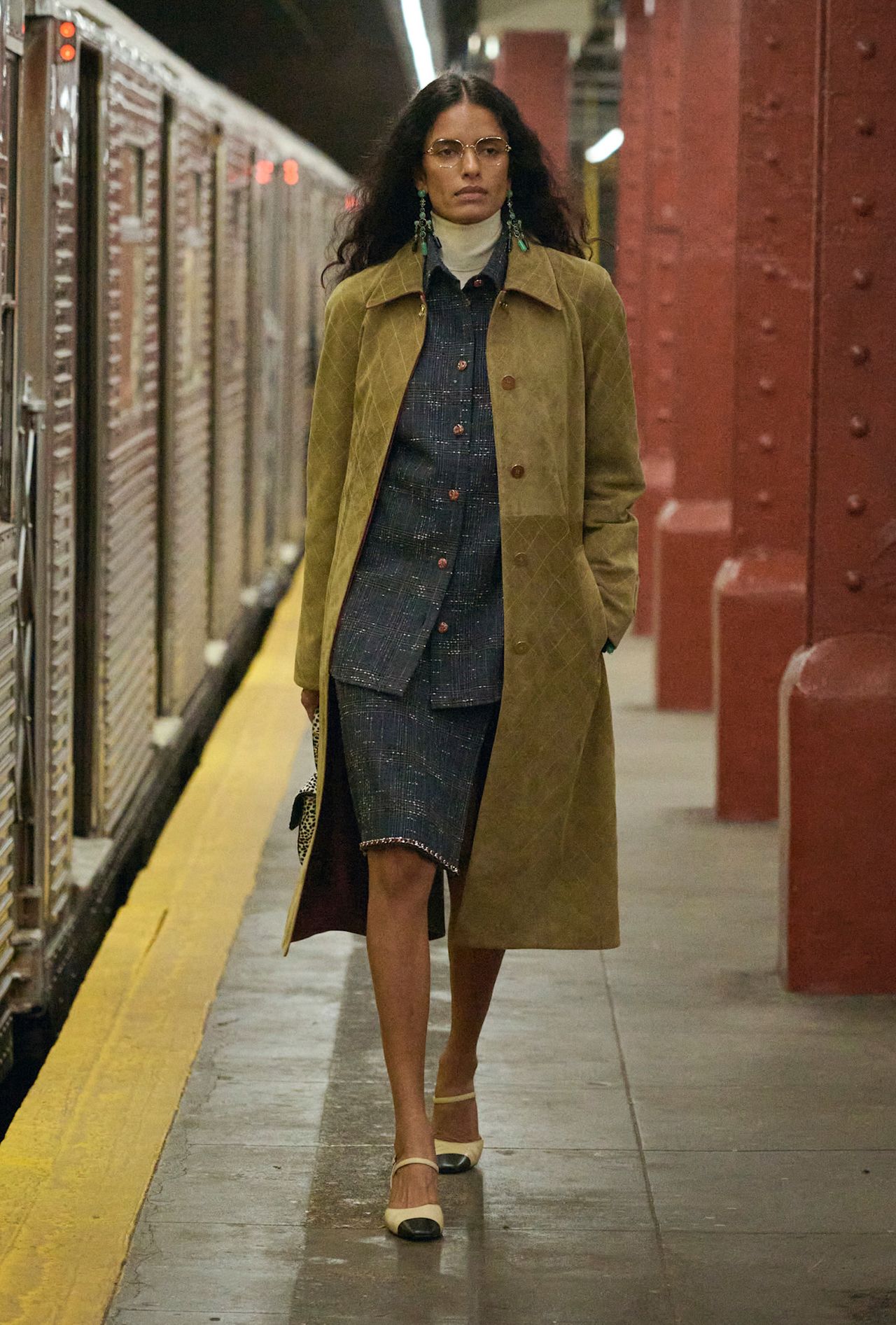 Layering staples for spring 2026 is shown in a photo from Chanel's runway show, with a model walking while wearing a tan suede coat layered on top of a navy blue button-down shirt with a white turtleneck peeking out underneath that's styled with a matching navy pencil skirt, cap-toe slingback heels, wireframe glasses, and green earrings