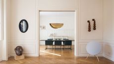 A white open room with hanging wooden sconces, a speaker on a tripod, a sculptural bust, and open doors leading to a dining space with a gold folded pendant light