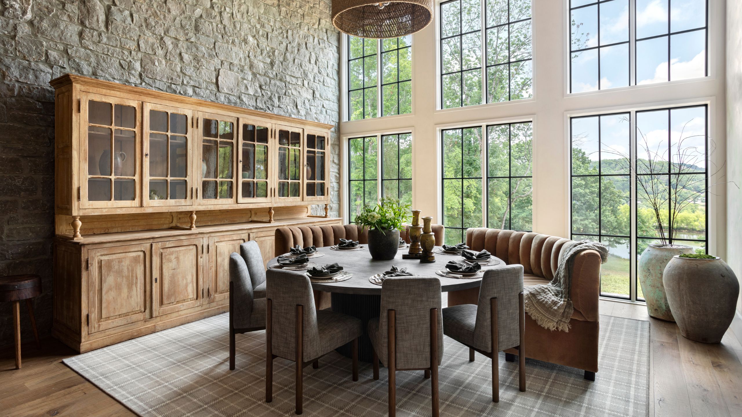 a large dining room with vaulted glass windows and a long stone wall with antique dresser and round table with leather bench seats