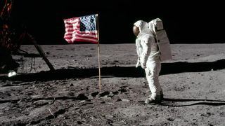 someone in a spacesuit on the surface of the moon stands, facing left, toward an American flag.