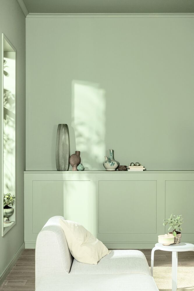 A modern living room drenched in a light sage green color, with light shining through the windows and hitting the wall. A built-in shelf holds various vases, along with a white sofa and side table.