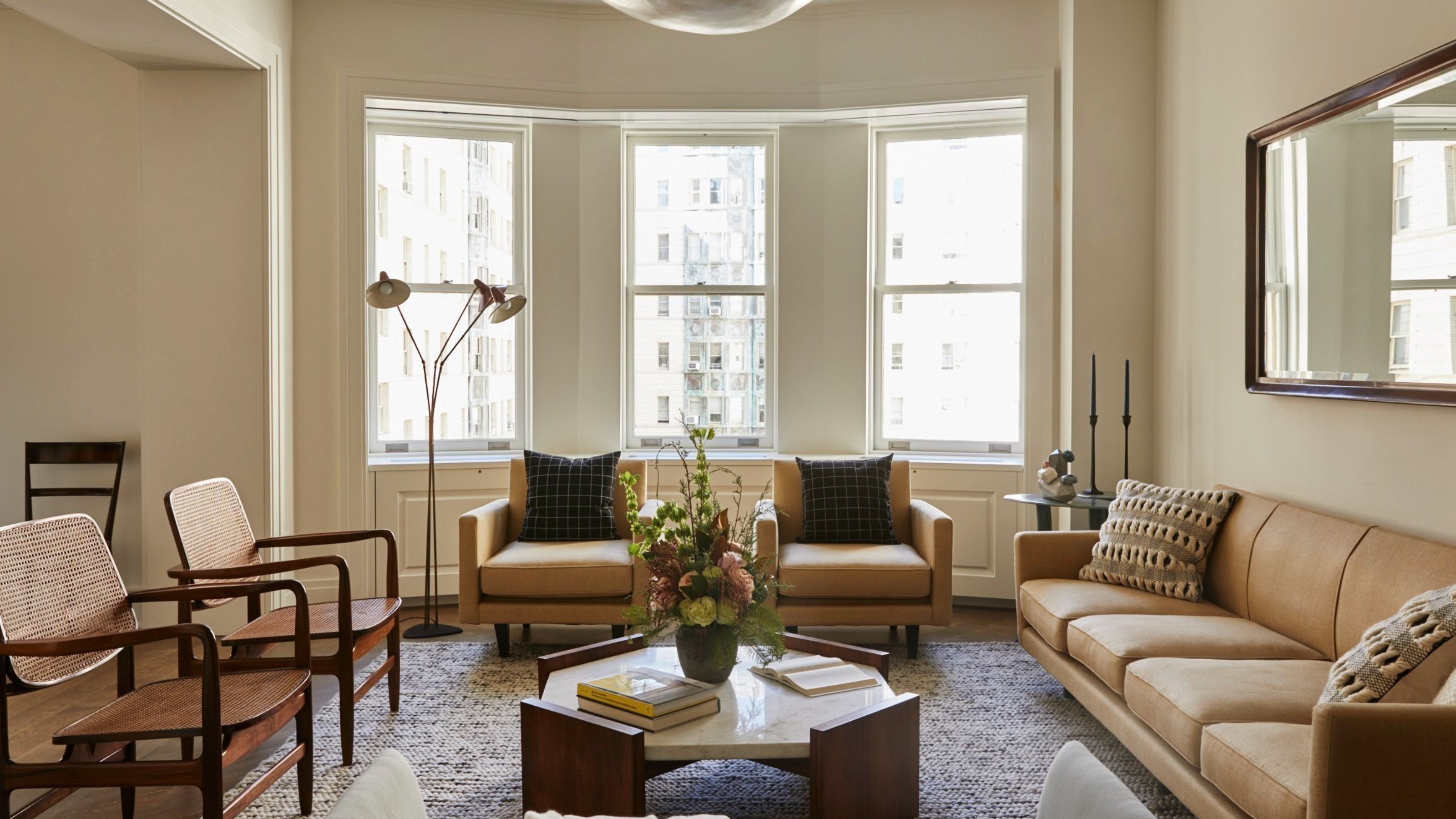 A neutral living room painting in a neutral paint color. There is a bay window at the back and a seating area made up of a tan sofa, two tan accent chairs, two wooden chairs, and a marble coffee table.