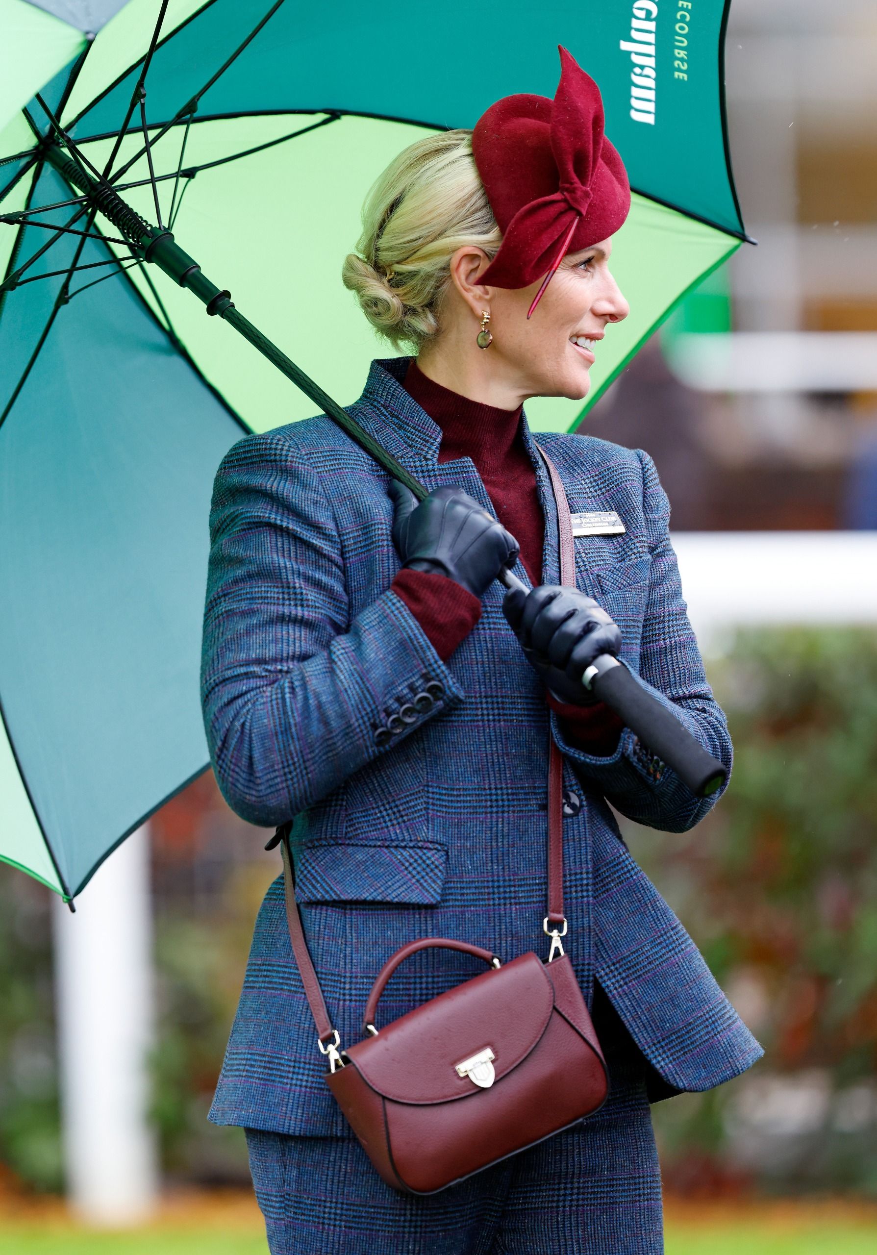 Zara Tindall wearing a gray suit holding an umbrella and a maroon bag
