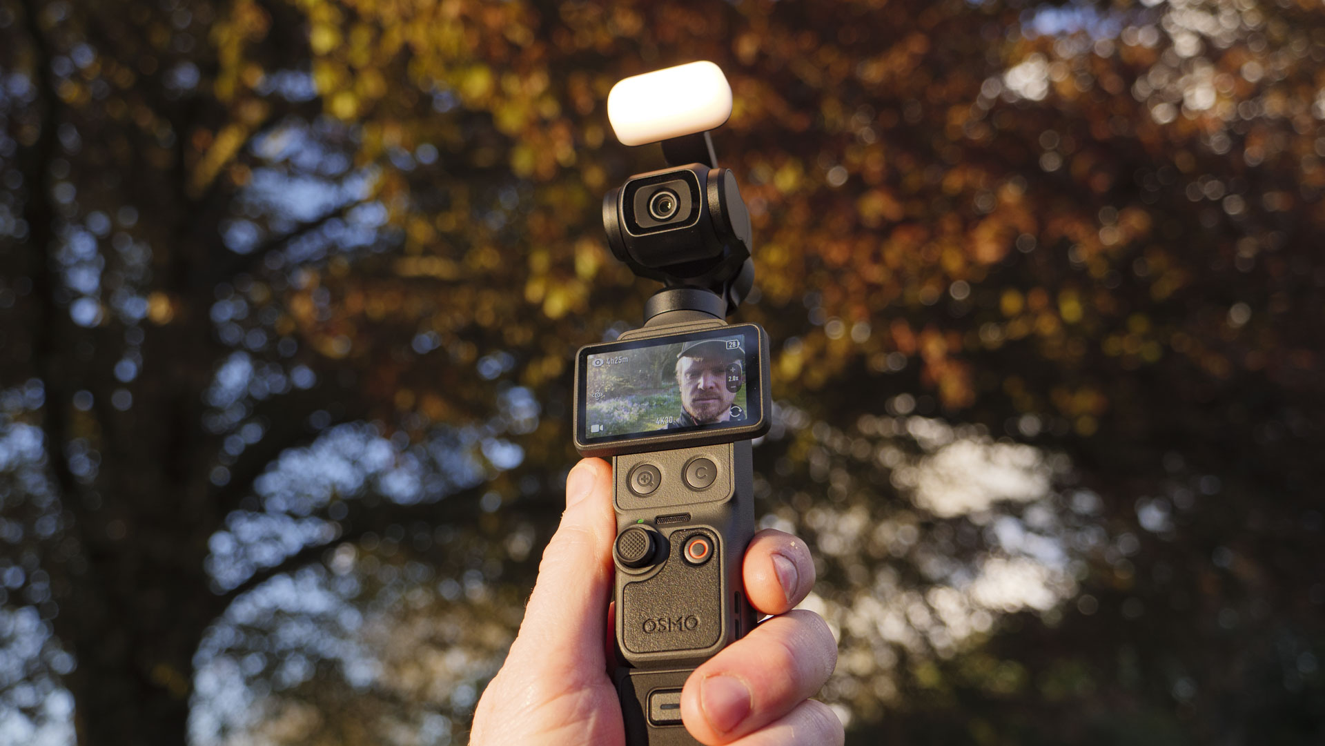 DJI Osmo Pocket 4 vlogging camera with natural background at golden hour, and the user in the frame of the screen