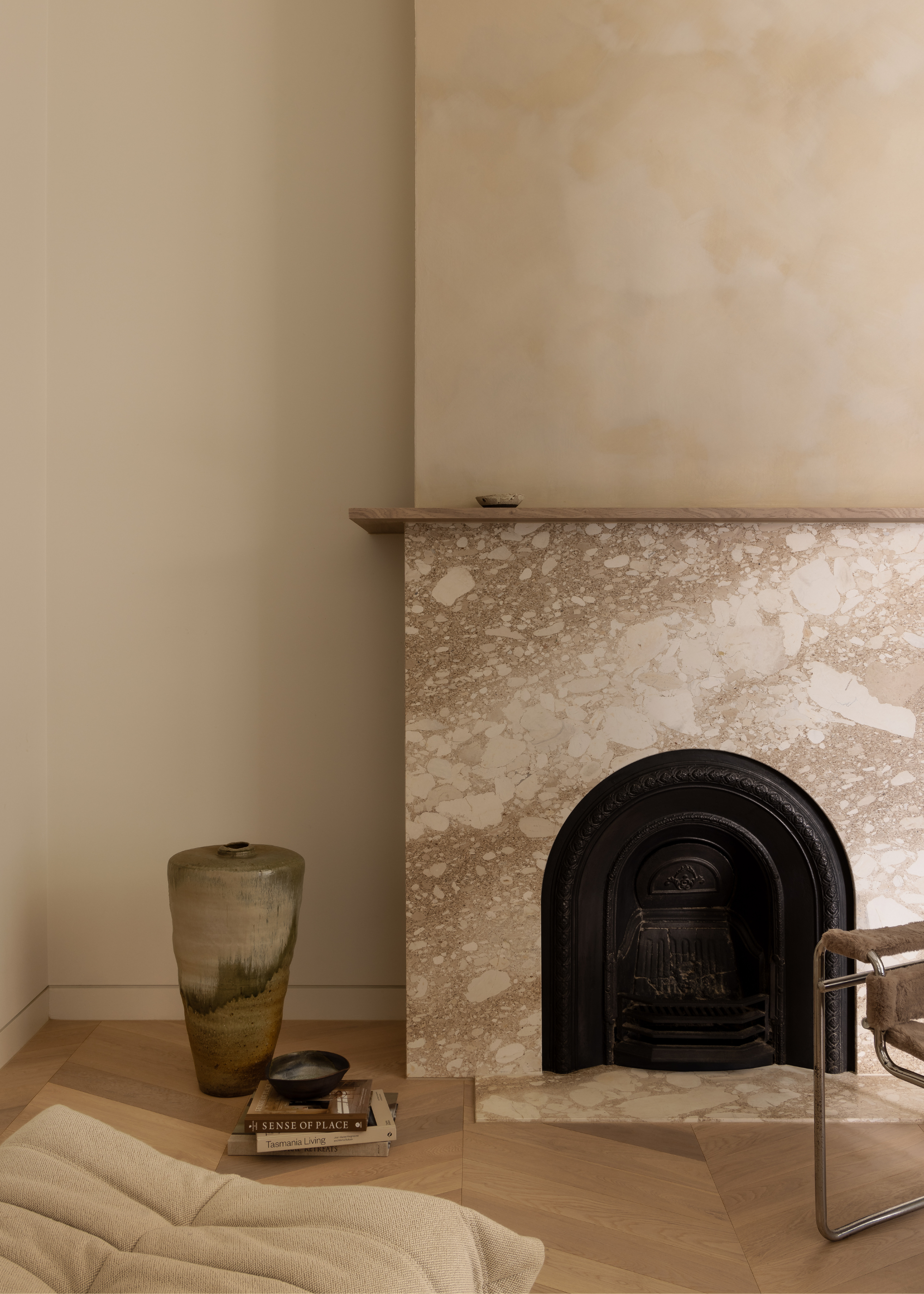 A minimalist fireplace with a stone mantel, a limewash accent wall, a vase, a stack of books, a beige Togo, and a tubular chair