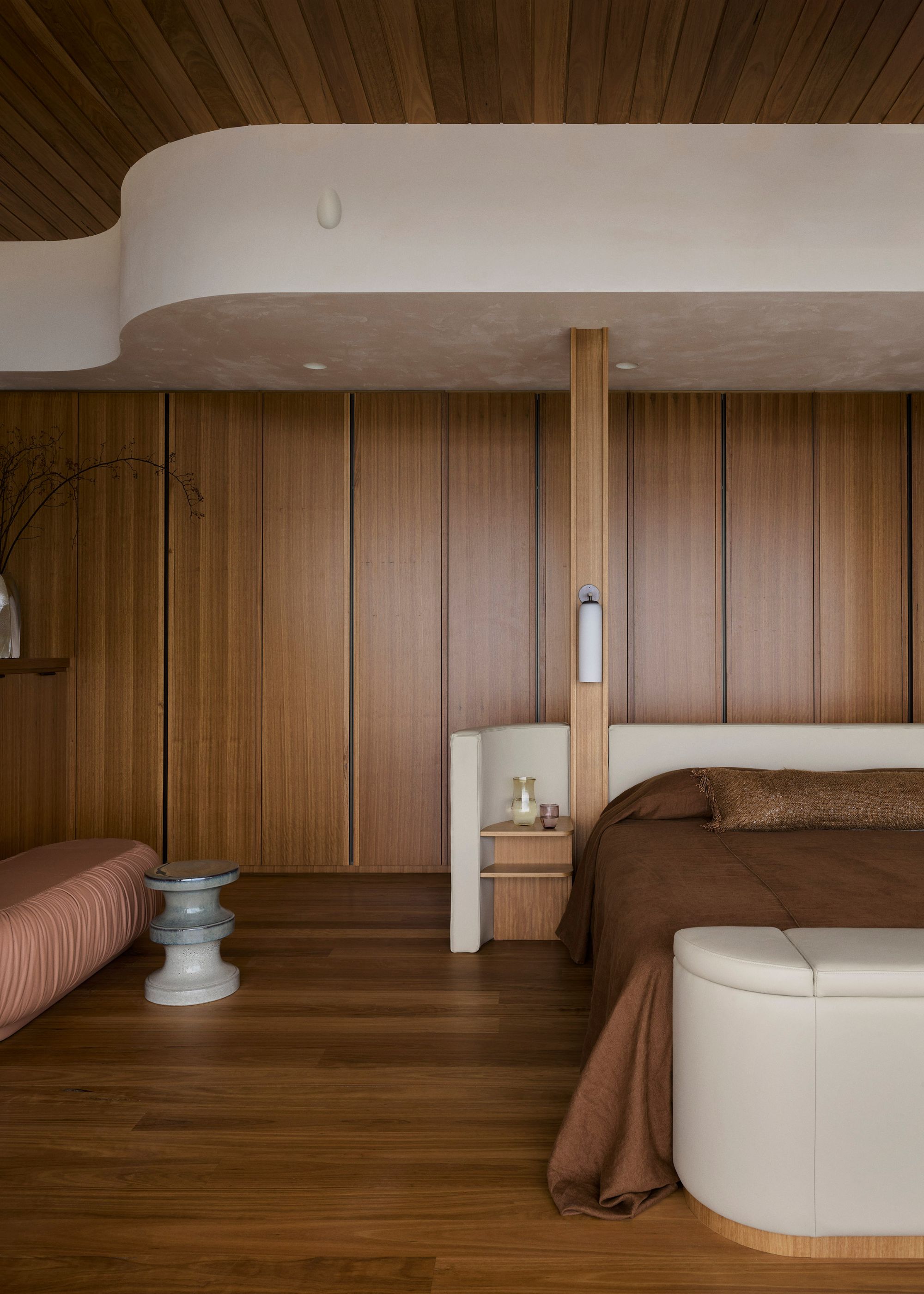 An earthy bedroom with wooden cupboards, a bed with brown bedding, a cream curved headboard, and attached nightstands, alongside a stacked stool