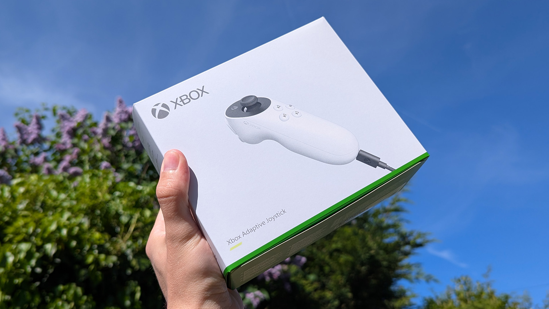 Xbox Adaptive Joystick in retail box, held in hand in front of a blue cloudy sky