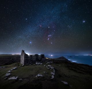 night sky with orion and the pleiades shine above castle ruins by the coast.