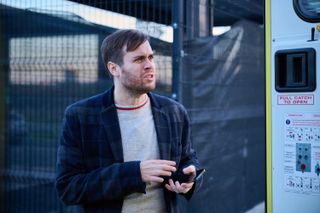 Spencer (Nathan Foad) stands outside the ambulance looking inward, wearing a blue-and-grey check jacket and a grey jumper, clutching his phone and with a look of mild disgust on his face