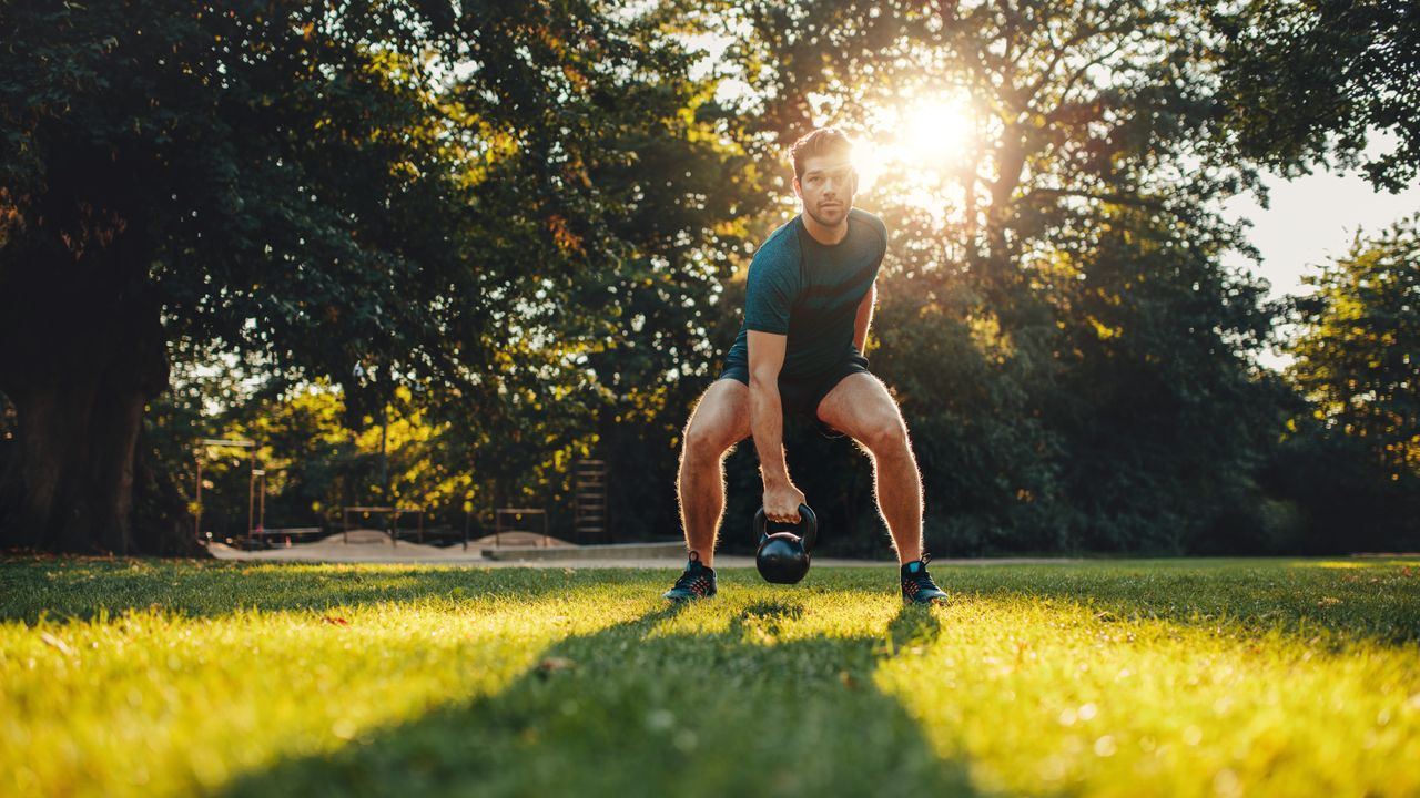 A man in shorts, t-shirt and sneakers exercises with a kettlebell outside. He&#039;s squatting down, knees bent, right arm extended and grasping a kettlebell between his legs. Underneath his feet is short-trimmed grass and behind him there are lots of leafy trees.