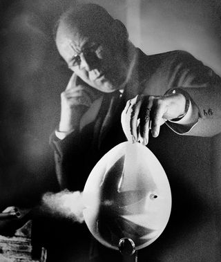 A bullet from a .22 caliber bullet starts to burst from side of a balloon held by Dr. Harold E. Edgerton in his Stroboscopic Light Laboratory at MIT in Cambridge, Mass., Feb. 19, 1959. An electronic photo flash invented and developed by Dr. Edgerton stopped the motion of bullet with a ½,000,000th of a second flash. Flash and camera were operated by sound of explosion picked up by microphone just below the balloon.